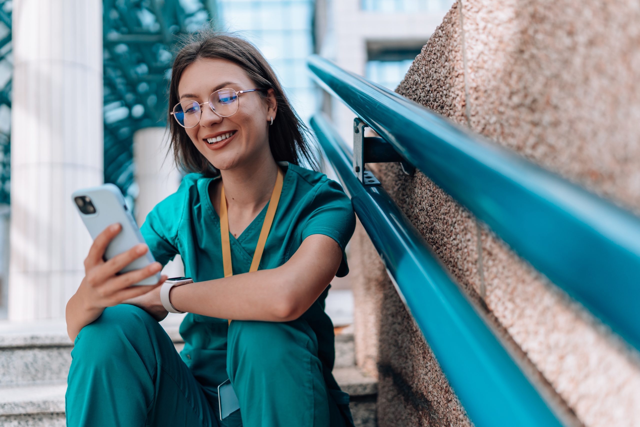 Young female nurse sitting on stairs in her scrubs, while cheerfully smiling and scrolling through social media Reapplying to medical school